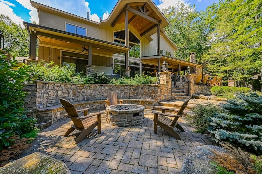 Large modern house with stone patio, fire pit, and Adirondack chairs, surrounded by nature.